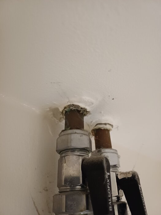 Två rör med ventiler tränger genom väggen, omgivet av smutsig isolering. Misstänkt asbest i ett badrum, lägenhet byggd kring 1950.