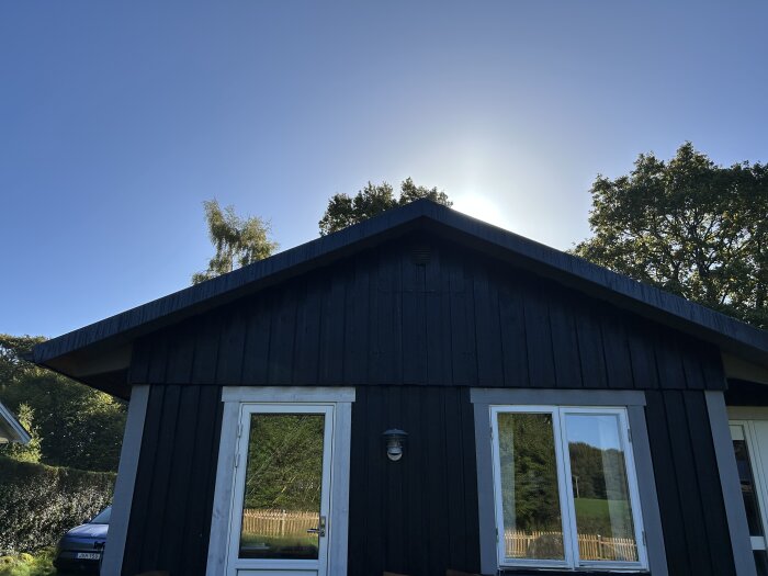 Svartmålat hus i trä med två vita fönster och en dörr under klar himmel; solens strålar syns ovanför taket.