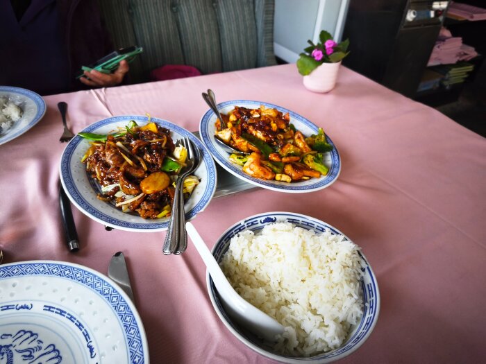 Asiatisk måltid på ett bord med två tallrikar kött och grönsaker, samt en skål med ris och en rosa bordsduk.