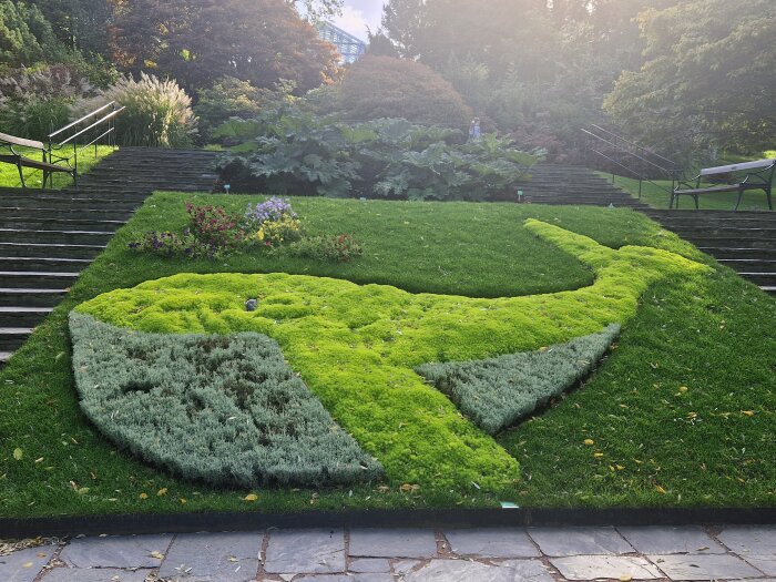 Grönskande växtinstallation formad som en val i en park med trappor på båda sidor och bänkar i bakgrunden.