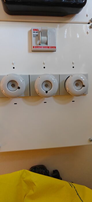 Tre vita gängade huvar på rad på en panel under en strömbrytare märkt "GARO HB 380", delvis skymda av en gul duk nedtill.