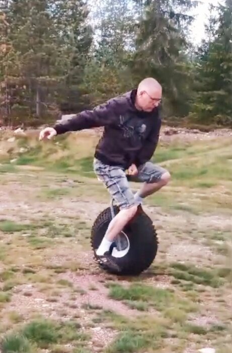 Man balanserar på ett enhjuligt fordon med ett stort terrängdäck utomhus på en gräsyta omgiven av träd.