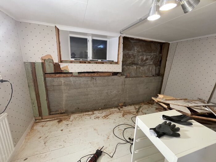 Källarrum under renovering med blottad naturstensgrund och halvnedriven vägg. Verktyg och handskar syns på golvet och ett bord.