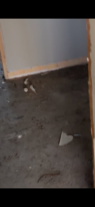 En byggarbetsplats med delvis färdig vägg, smutsigt golv och byggavfall, inklusive träbitar och gipsfragment.