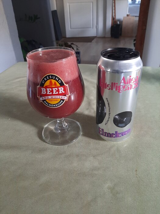 Ett glas med röd skummande dryck märkt "Öresund Beer" bredvid en silverfärgad burk med texten "Alien Intelligence Elm Eleven" på grön duk.