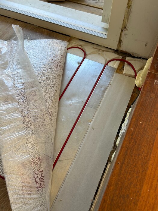 Elektrisk golvvärmeslinga synlig under plastlaminat och Kährs golvunderlag, vid en dörröppning.