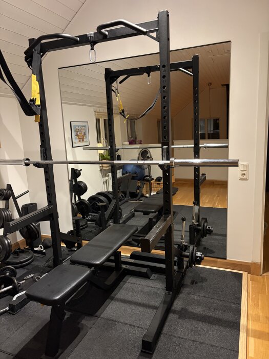 Hemmagym med ett ställ för styrketräning, bänk och vikter i ett rum med spegel och trägolv.