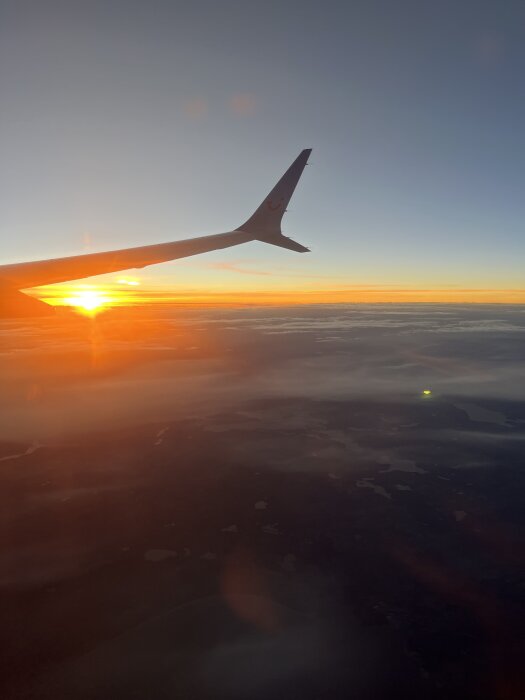 Utsikt från ett flygplansfönster med vinge synlig, solnedgång över molnen och landskap nedanför.