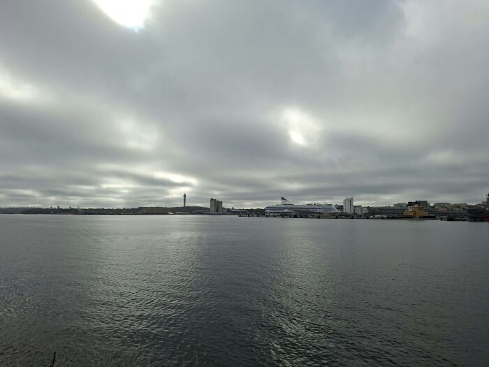 Vy över en hamn med en stor färja vid kajen, byggnader och en hög mast syns i bakgrunden under en mulen himmel.