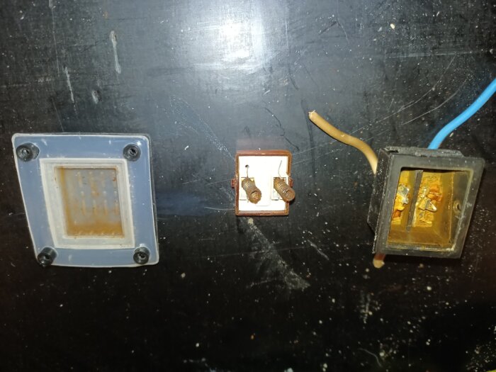 Tre delar av ett elektriskt uttag. En ram, en panel med skruvar och en elektrisk del med kablar. Tecken på rost syns.