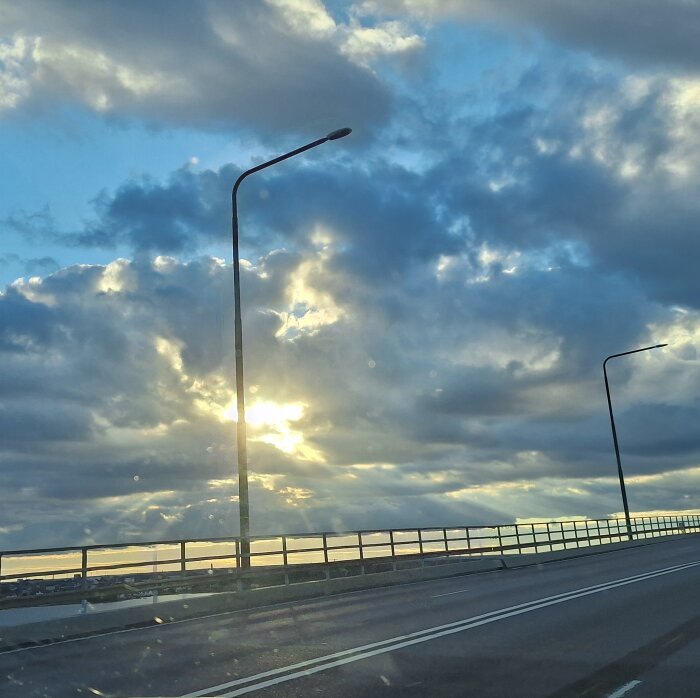 Solnedgång över en bro med molnig himmel och lyktstolpar som syns mot den ljusa horisonten.