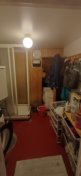 Tvättstuga med röd matta, duschkabin, vita hyllor, klocka på väggen och flera upphängda jackor och påsar. Lampan är tänd i taket.