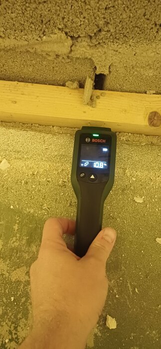Hand håller en fuktmätare mot en träregel med gips och mineralull runt omkring, visa 11% fuktighet.