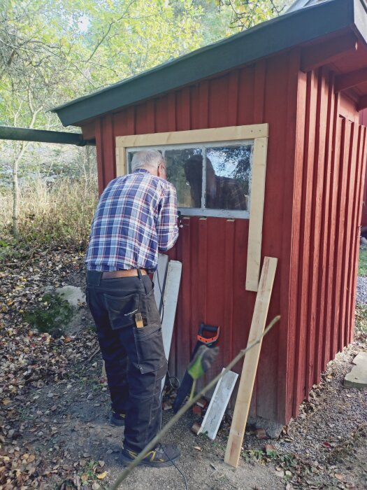 Man använder en skruvdragare på röd träbyggnad; verktyg och träbitar syns bredvid.