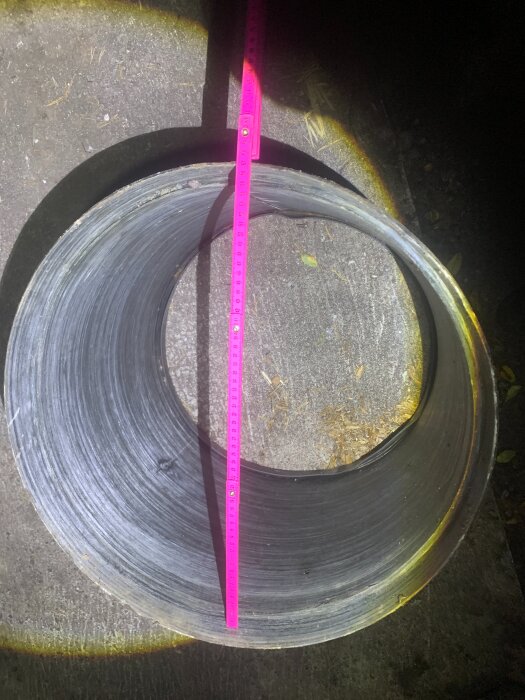 Metallisk förhöjningsring placerad på marken med en rosa måttstock över öppningen, avsedd för användning med en fördelningsbrunn.