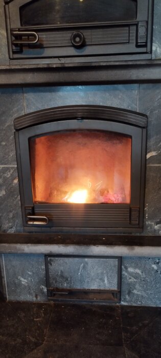 Kamin med eldstad som brinner, omgiven av mörkt stengods eller kakel. Flammor syns genom glasluckan.