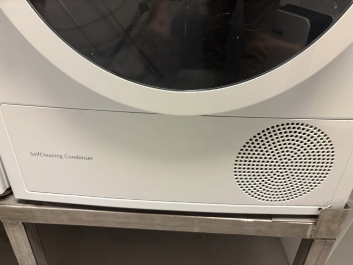 Lucka med texten "SelfCleaning Condenser" på vit hushållsapparat, med cirkulära ventilationshål.
