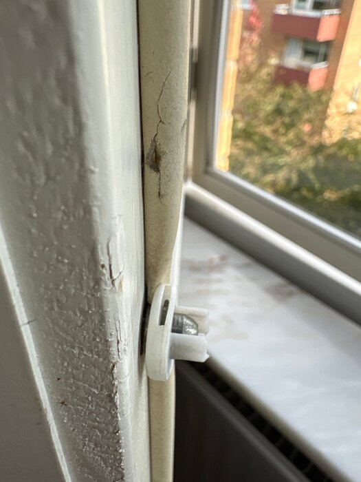Plastdel i fönsterkarm som inte låser ihop inner- och ytterfönster, med sprucken träram i bakgrunden.