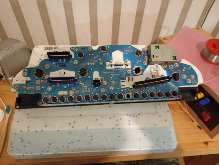 Instrumentpanelens kretskort från en bil med synliga lödpunkter och komponenter, placerad på ett bord med verktyg i närheten.