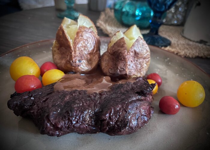 Grillad entrecote med pepparsås, två bakade potatisar och färgglada egenodlade tomater på en tallrik.