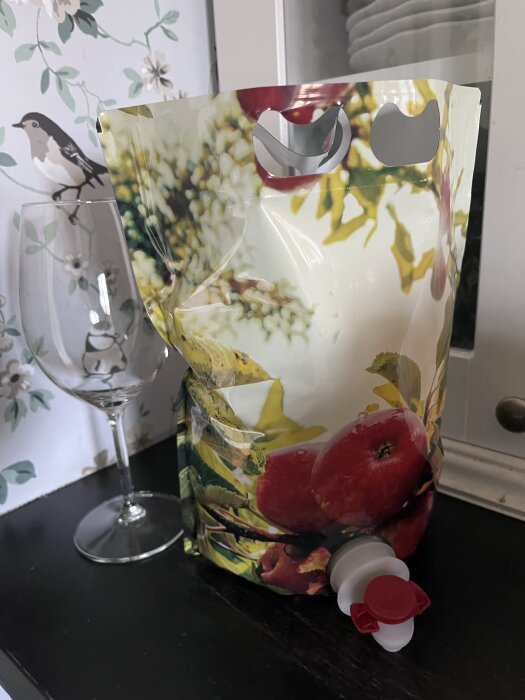 Förpackning med äppelmust på en hylla bredvid ett vinglas, dekor med äpplen och blommor på förpackningen.