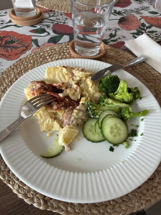 Tallrik med potatisgratäng, broccolibitar och gurkskivor. Glas med vatten på bord med blommigt tyg.