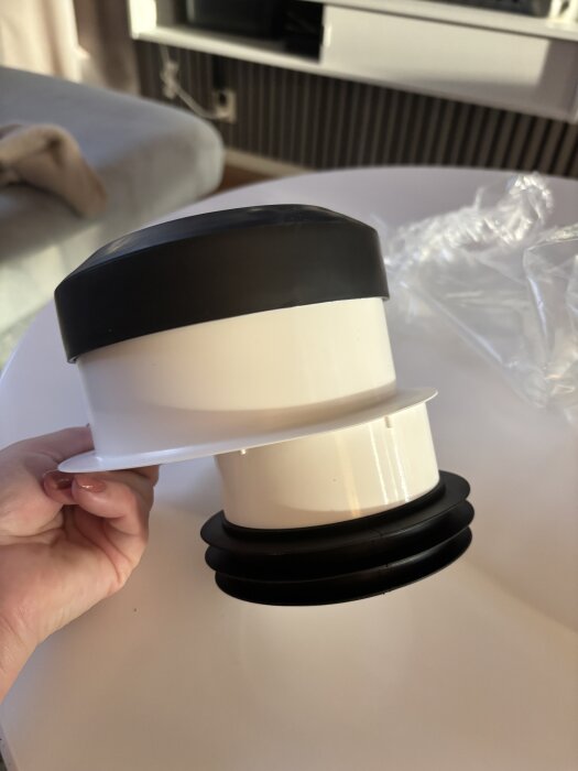 Vit och svart toalettanslutning i plast med en hand som håller i den, ligger på ett bord framför en soffa.