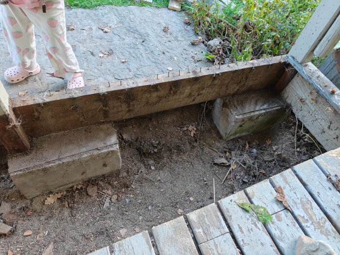Betongplintar och jord vid tidigare altan, träbjälke med spik ovanpå plintar. Ett barn står nära i områdets övre vänstra del.