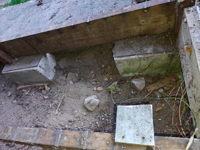 Betongblock och träplankor under konstruktion, omgivna av jord och stenar, med viss grön växtlighet kring strukturen.