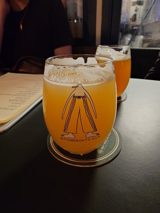 Ett glas med öl märkt "Stigbergets Fot", stående på ett underlägg, på ett bord i en bar eller restaurangmiljö.