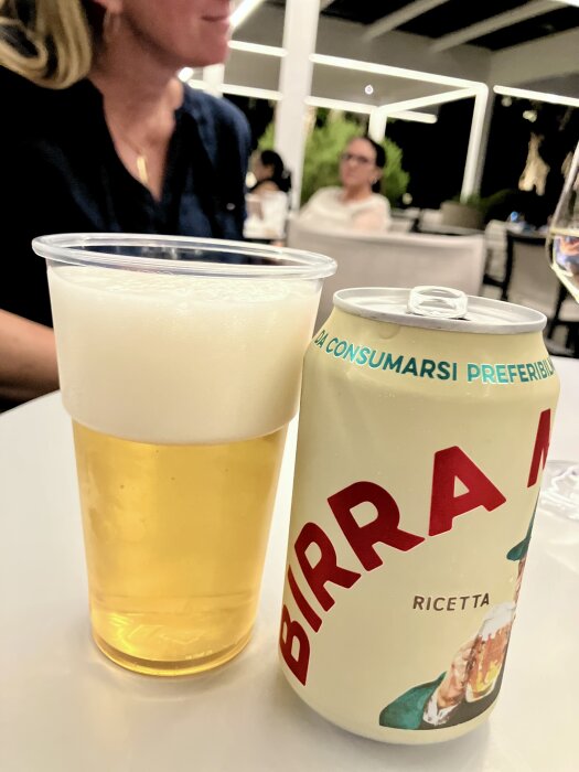 Öl i plastglas bredvid en burk med etiketten "Birra Moretti", personer i bakgrunden i restaurangmiljö.