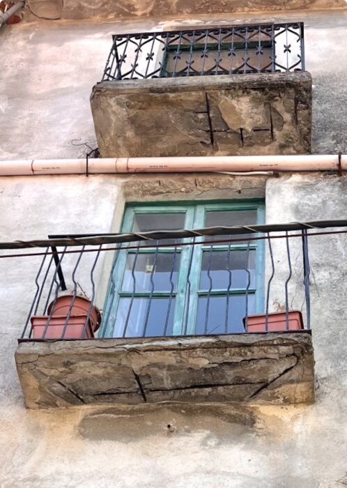 Sliten balkong med sprucken betong och blottad armering, utsmyckad med metallräcke och blomkrukor, belägen vid ett hus i södra Italien.