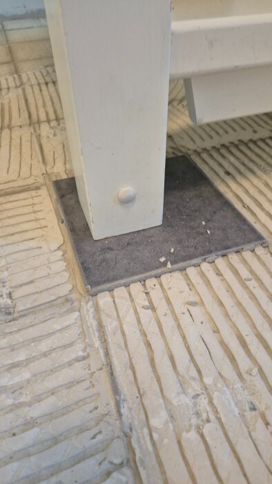 Närbild av ett golv där en brun klinkerplatta ligger delvis under en vit trappa, omgiven av ett rutigt mönster från borttagen klinker.