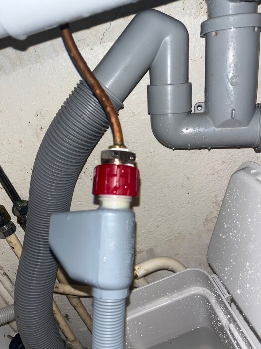 Kopparrör med gängor anslutet till en grå plaströrkoppling och en röd mutter under diskbänken, del av diskmaskinsinstallation.