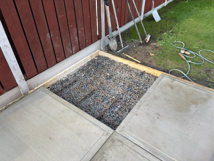Grusunderlag med armeringsnät förberett för betonggjutning bredvid en nygjuten betongplatta invid en röd trästaket. Verktyg och slangar syns.