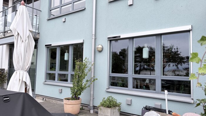 Nyinstallerad markis med vertikal screen ovanför fönster på ett hus, omgiven av växter i krukor på en uteplats.