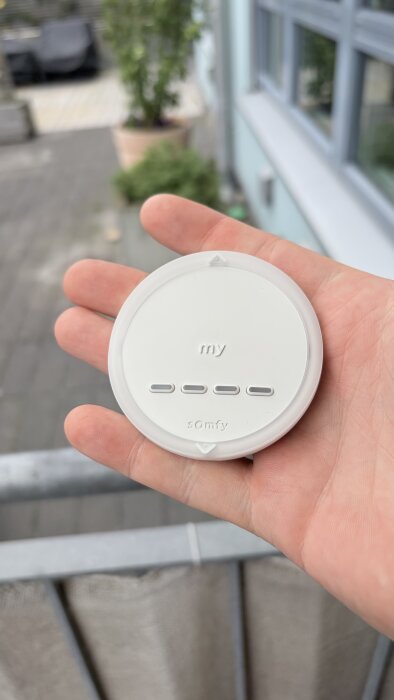 En hand håller en rund fjärrkontroll för markis av märket Somfy med symboler och text "my." Bakgrunden visar ett utomhusområde.