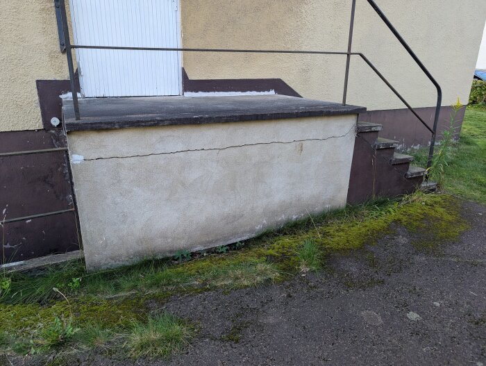 Spricka i murad vägg vid källaringång med trappa och räcke, marken är täckt med mossa; potentiell strukturell skada.