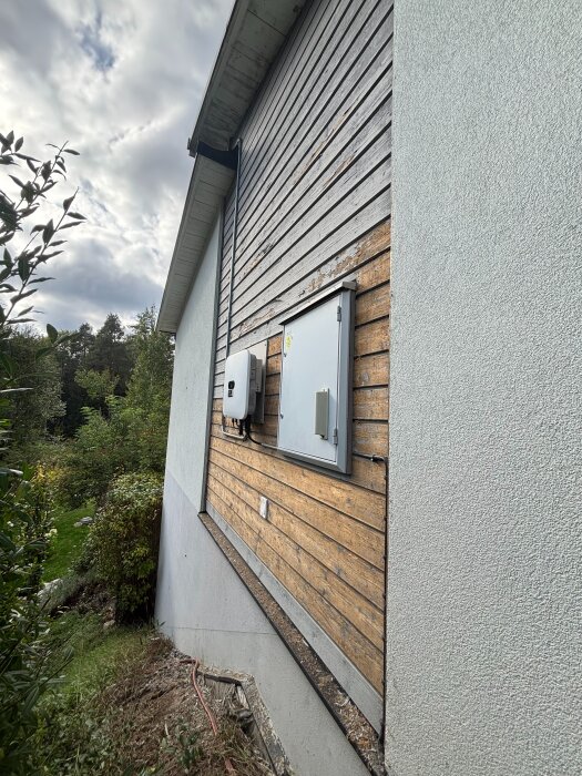 Vägg med träpanel och puts, flagnad färg på träet och två elboxar. Marken är ojämn och det finns buskar och gräs nära väggen.