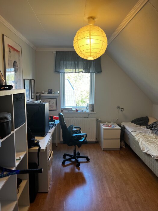 Rum med snedtak, hålkärlslist runt taket, skrivbord, kontorsstol, säng och stor papperstake.