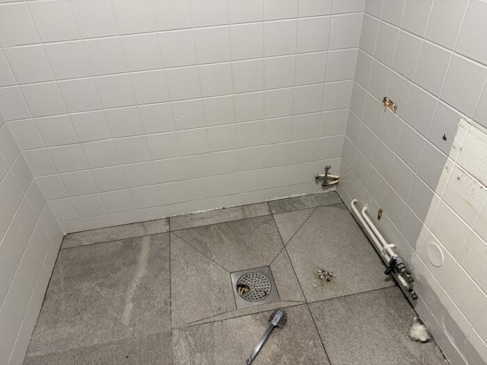 Tomt badrumshörn med grått kakel golv, vita väggplattor, och synliga rör där ett badkar tidigare stod, dräneringsgaller på golvet.