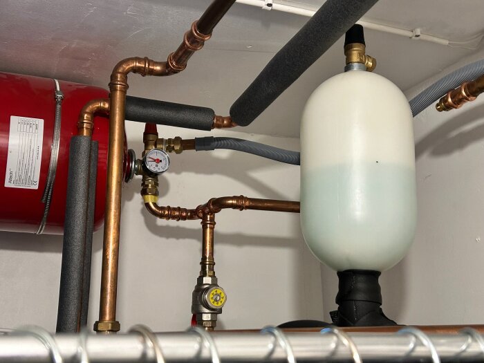 Värmesystem med kopparrör, en tryckmätare och ett halvtfyllt plastkärl. Synliga fuktfläckar och röd cistern i bakgrunden.
