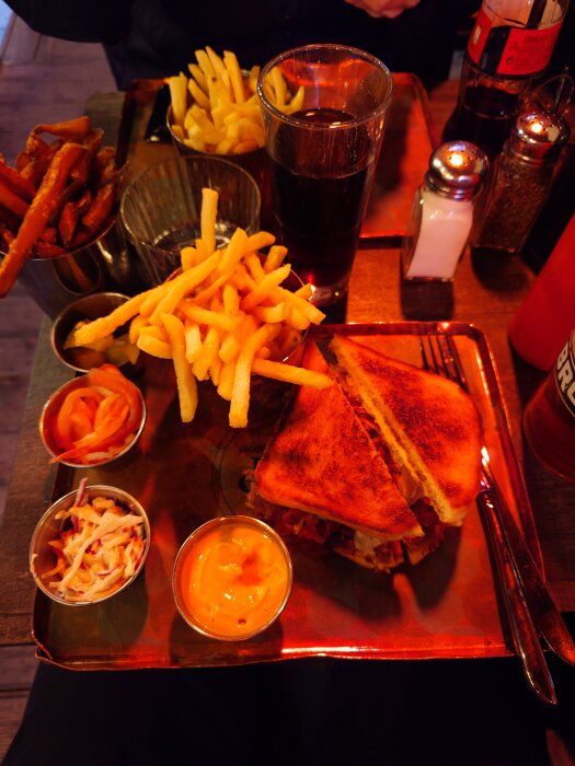 En Pappastrami-smörgås på ett fat med tillbehör, inklusive pommes frites, dippsåser och en dryck i ett glas.