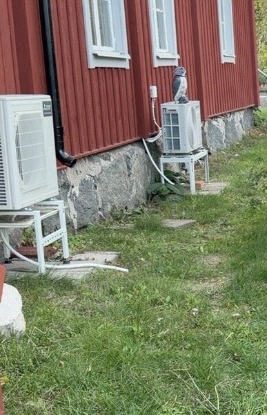 Röd husfasad med naturstensgrund, två luftkonditioneringsenheter monterade utanför, grön gräsmatta i förgrunden.