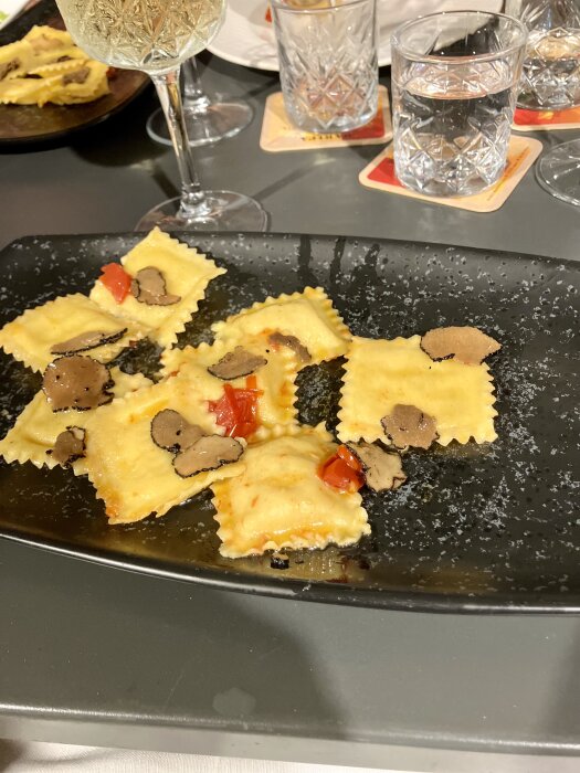 Ravioli med scampi och tryffel på svart tallrik med röd paprika dekoration, serverad med vinglas och glas med vatten i bakgrunden.
