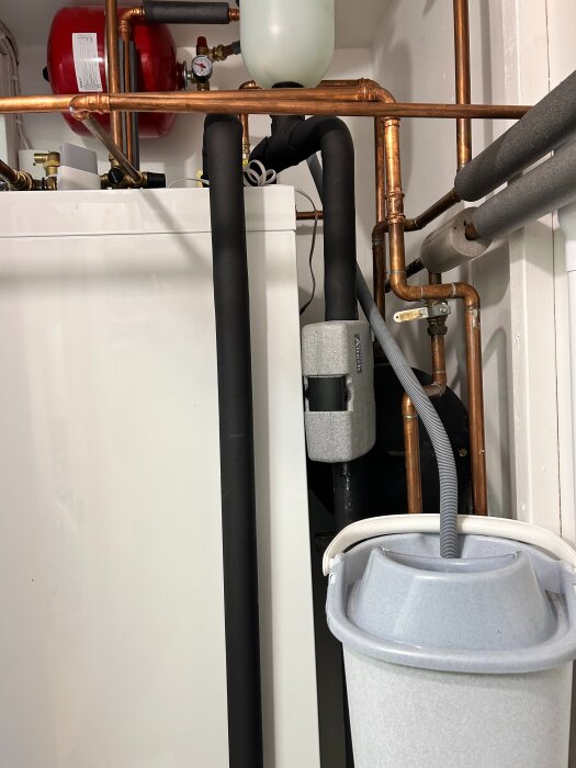 Vattenvärmepanna med kopparrör och svart isolering i en teknisk installation. Hink med slang under rörsystemet.