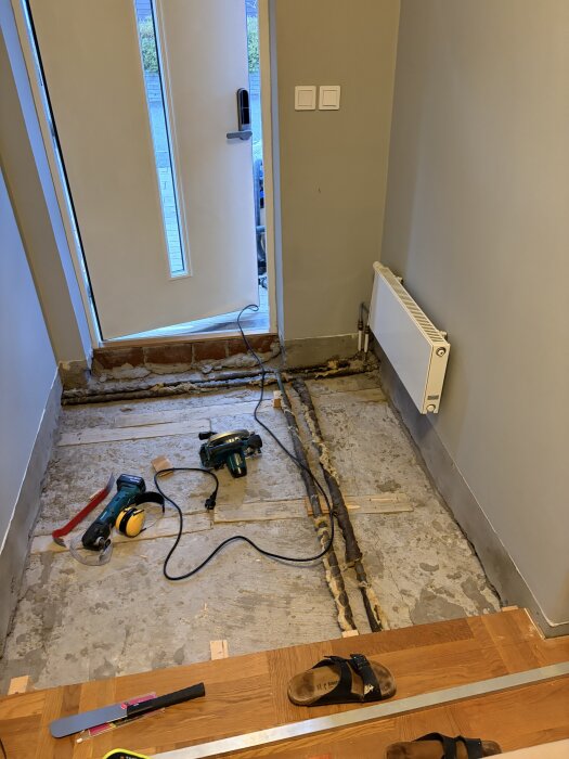 Renovering av golv, med exponerat betongunderlag. Verktyg som spårfräs och säkerhetsutrustning är utplacerade på golvet nära en öppen dörr.