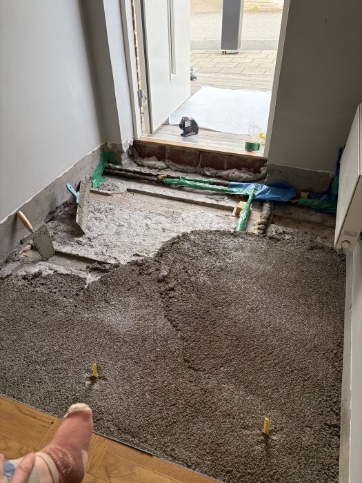 Renoveringsarbete med exponerad golvstruktur och cement, synlig dörröppning i förgrunden. Verktyg och material runt arbetsområdet.