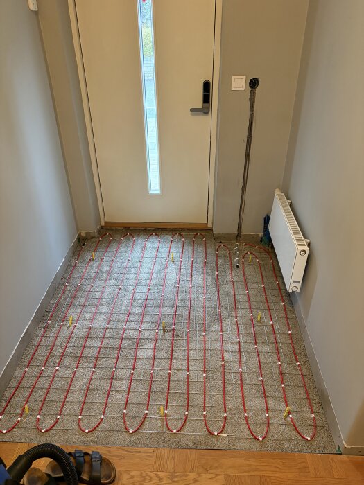 Golvvärmeslingor installerade på ett betonggolv framför en dörr, kablarna är organiserade i jämna rader, redo för flytspackling och klinkerläggning.