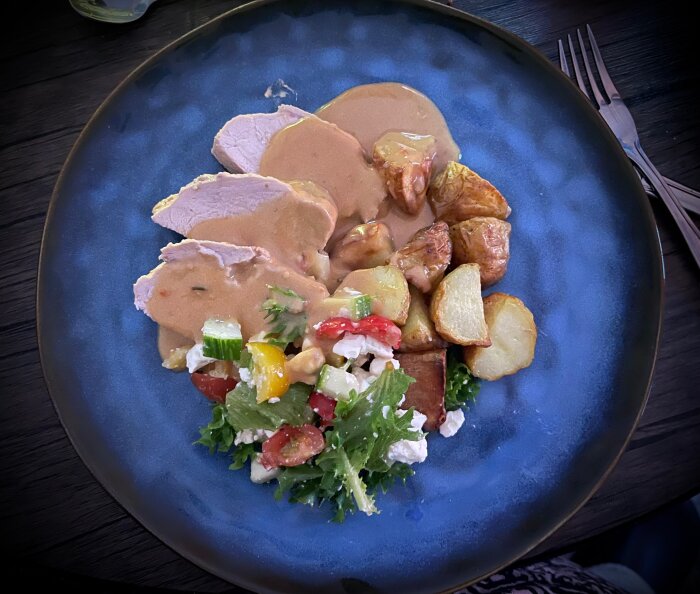 Fläskytterfilé med sås, serverad med ugnsstekt potatis och färgglad sallad på en blå tallrik.